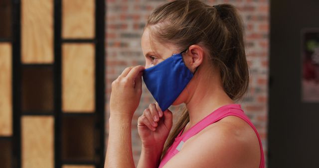 Woman wearing a blue face mask and ponytail adjusting her mask while at the gym. Perfect for concepts related to health and safety measures during workouts, COVID-19 precautions, and fitness environments.