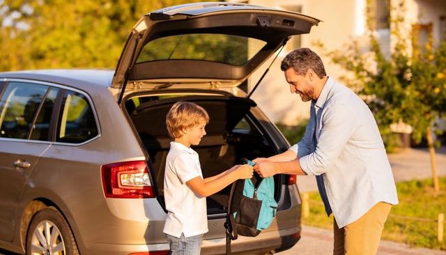 Father passing backpack to son at car trunk in suburban driveway morning routine