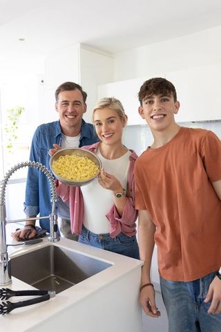 Happy family cooking together in bright modern kitchen holding colander of pasta smiling