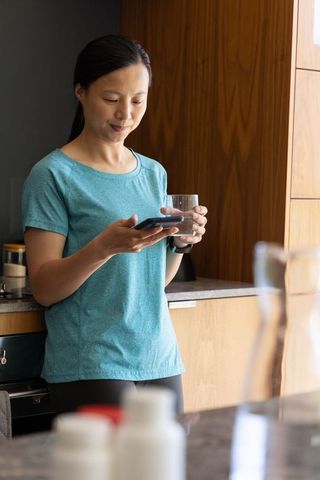 Asian woman checking smartphone while holding glass of water in modern kitchen
