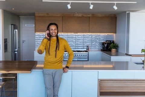African american man talking on phone leaning on modern white kitchen island smiling lifestyle portr
