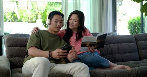 Asian couple relaxing on sofa sharing music and book in bright modern living room