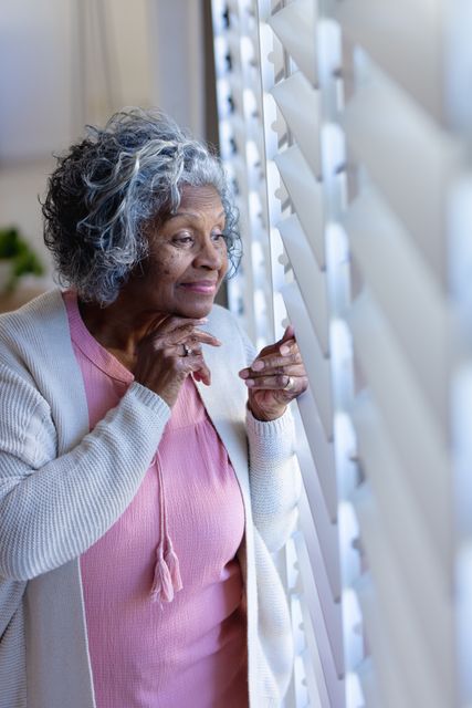 Vertical image of senior african american woman at home looking out of window smiling, copy space. Retirement, senior health, happiness, inclusivity and senior lifestyle concept.