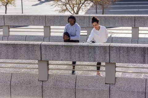 Two women stretching on urban concrete barrier wearing hoodies activewear morning workout
