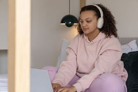 African american woman typing on laptop and listening with headphones in cozy bedroom