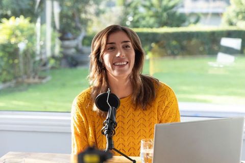Young woman recording podcast in sunlit home studio with microphone laptop and garden view