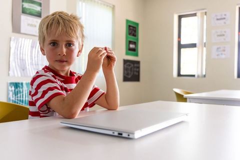 Young boy in red striped shirt using laptop in classroom holding small object, focused