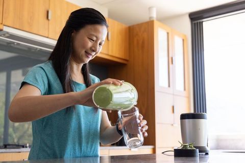 Asian woman pouring green smoothie into glass in modern kitchen promoting healthy living