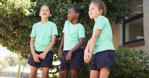 Three preteen girls in light green shirts and navy shorts standing looking up at school