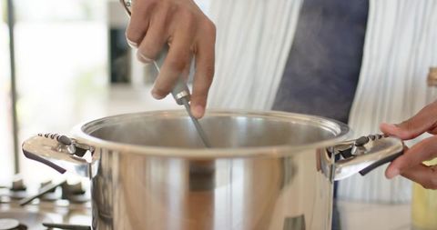 Closeup man stirring stainless steel stock pot on gas stove with rising steam in home kitchen