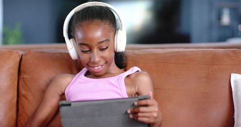 Preteen african american girl listening with headphones using tablet on brown leather sofa