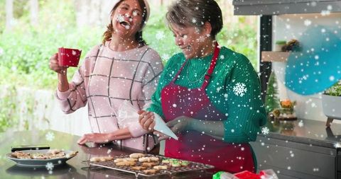 Two women decorating holiday cookies in kitchen with festive snowfall overlay