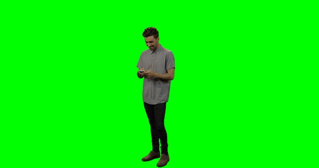 Man wearing casual clothing standing and texting on smartphone against green screen background. Suitable for video editing, presentations, and graphic design projects where green screen technique is needed for background removal or replacement. Can be used to depict themes of modern communication, technology, or daily activities.