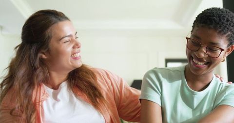 Two diverse women laughing together on couch in bright cozy room wearing casual tees and glasses