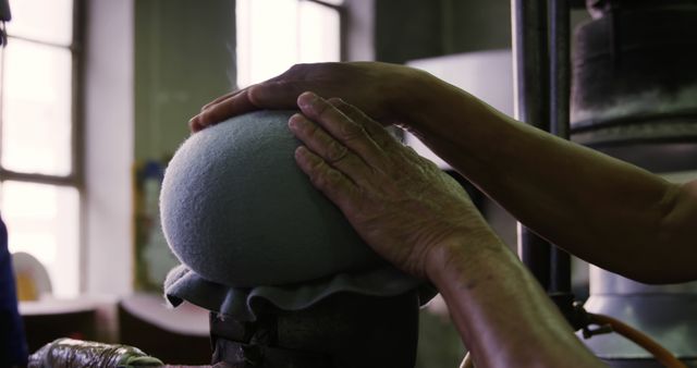 Craftsman is shaping a grey felt hat using a traditional steam machine in a workshop. This image can be used to illustrate articles on traditional crafts, handmade goods, and artisan skills. It is ideal for content related to skilled trades, artisanal products, and factory work.