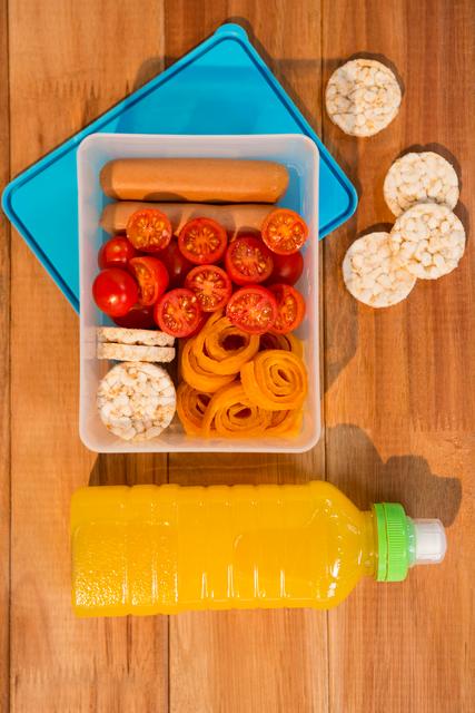 This image shows a variety of healthy snacks including cherry tomatoes, rice cakes, carrot spirals, and a hot dog in a lunchbox, accompanied by a bottle of orange juice. Ideal for illustrating concepts related to healthy eating, meal preparation, balanced diets, and nutritious snacks. Suitable for use in articles, blogs, and advertisements promoting healthy lifestyles and meal planning.