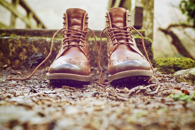 Vintage leather boots with laces resting outdoors on rustic ground with leaves and moss. Perfect for themes related to hiking, adventure, nature, rugged lifestyle, and outdoor footwear. Great for advertisements, blog posts, and articles about outdoor activities, exploring, and durable footwear.