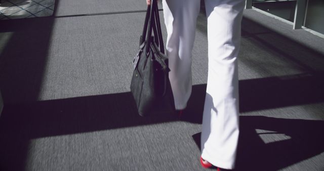 Businesswoman walking in an office hallway, carrying a black handbag while wearing white pants and red heels. Image can be used in articles about corporate work life, professionalism, office attire, women's workplace fashion, and corporate environments.