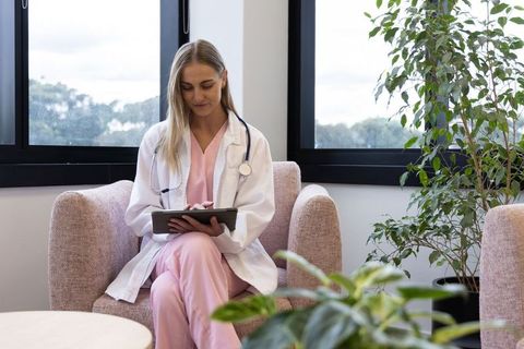 Female clinician using tablet in calm clinic lounge wearing pink scrubs and white coat