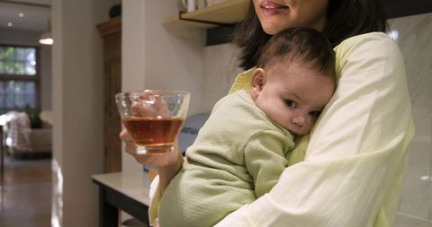Mother Cradling Baby While Holding Tea in Warm Kitchen Environment