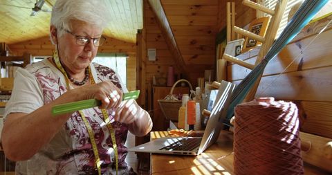 Senior woman weaving and working on laptop in sunlit workshop