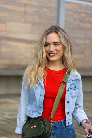 Young woman smiling in denim jacket and red top with olive crossbody bag in city plaza