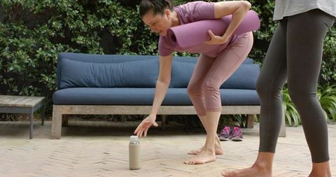 Diverse senior women practicing outdoor yoga holding mat, reaching for water bottle on patio