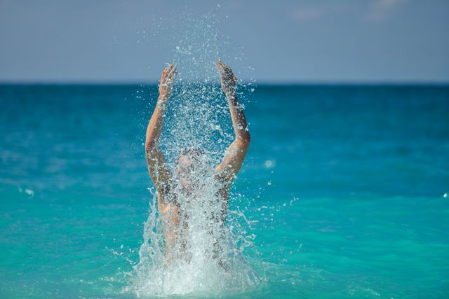 Person enjoying a fun time in a tropical ocean, splashing water with hands. Perfect for travel blogs, summer vacation advertisements, lifestyle magazines, and health and wellness promotions. Highlights joy and relaxation in a serene outdoor environment.