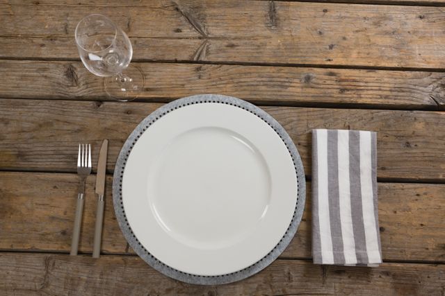 Overhead view of a rustic table setting featuring an empty plate, wine glass, striped napkin, fork, and butter knife on a wooden table. Ideal for use in articles or advertisements related to dining, hospitality, food presentation, or rustic-themed events.