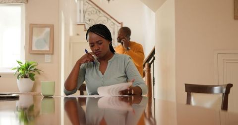 Focused woman reviewing documents while man talks on phone at home kitchen