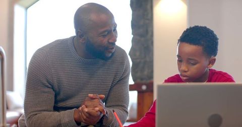 African american father teaching son homework using laptop in bright living room