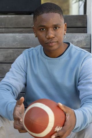 African american teen athlete holding football on wooden steps wearing blue sweatshirt