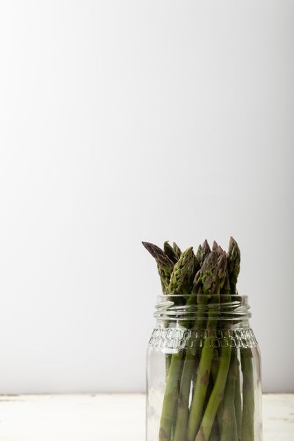 Fresh asparagus spears standing upright in a glass jar against a plain white background. Ideal for use in healthy eating promotions, organic food campaigns, nutrition blogs, and minimalist food photography. The image's clean and simple composition makes it perfect for advertisements, recipe books, and social media posts focused on fresh produce and clean eating.