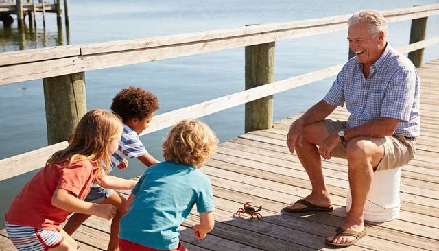 Grandfather smiling while kids crouching and watching crab on sunny wooden pier by water