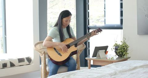 Young woman playing acoustic guitar in bright bedroom during online music lesson