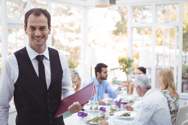 Portrait of smiling waiter holding menu while friends dining in background in restaurant