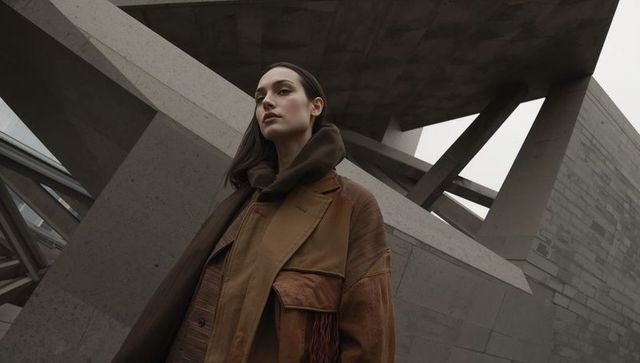 Low-angle fashion portrait showcasing oversized brown coat and scarf against brutalist concrete