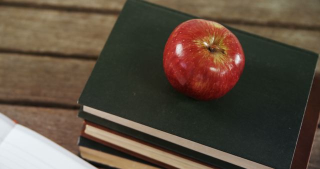 A red apple sits atop a stack of books on a wooden table, symbolizing education and healthy eating. Often associated with teachers and students, the apple represents a traditional gift for educators and a focus on academic success.