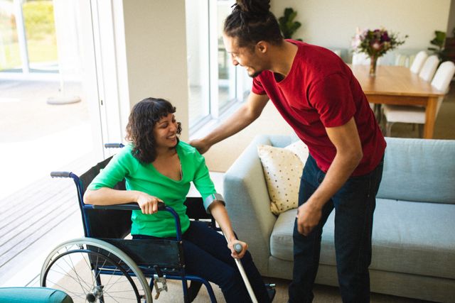 Smiling biracial man helping young woman getting up from wheelchair in living room at home. support, disability and love, unaltered.