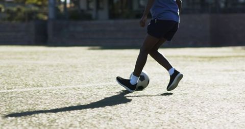 Sunlit teenager dribbling soccer ball on turf casting long shadow during practice