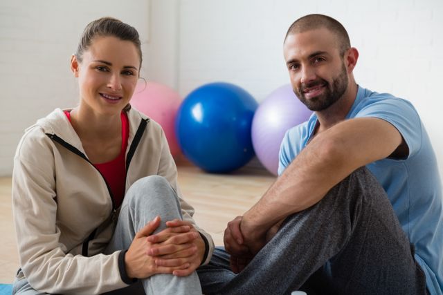 Female student and instructor sitting on floor in yoga studio, smiling and relaxing after a session. Ideal for use in fitness, wellness, and health-related content, promoting yoga classes, gym memberships, and active lifestyle campaigns.