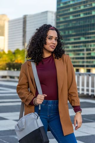 Young professional woman standing on urban rooftop wearing brown blazer maroon top shoulder bag