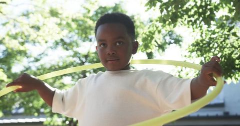 African american boy playing with neon hula hoop under sunlit backyard trees