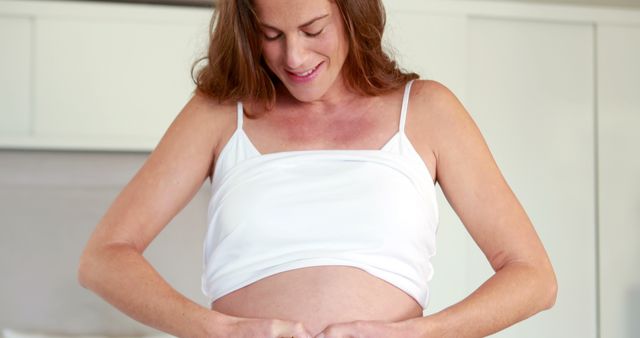 Expectant mother wearing white tank top smiling while cradling her belly in a comfortable home environment. Perfect for use in articles, blogs, and advertisements related to pregnancy, maternity care, prenatal health, parenting, and women's health.