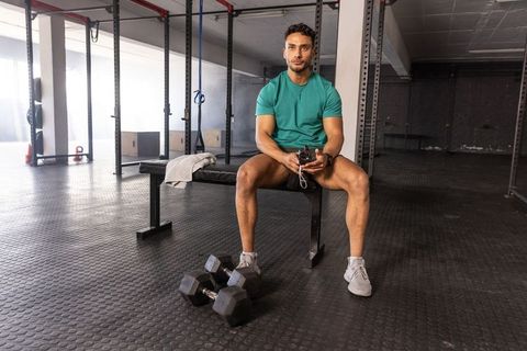 Fit man resting on bench holding stopwatch after intense strength workout in modern gym