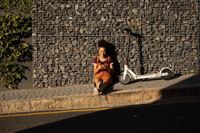 Front view of a happy biracial woman enjoying free time in a city on a sunny day, sitting on pavement, using smartphone, an electric scooter next to her.
