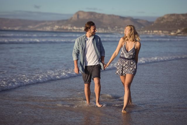 This image shows a happy couple holding hands while walking along the shore at a beach. The scene captures a romantic and carefree moment, perfect for use in travel brochures, romantic getaway advertisements, relationship blogs, or social media posts about love and vacations.