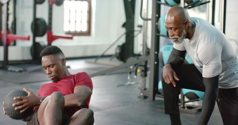 African american trainer coaching male client doing medicine ball core workout in gym