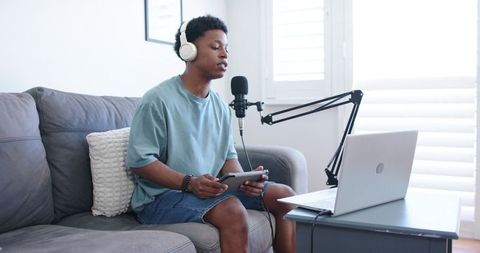 Young man podcasting from living room wearing headphones and using tablet