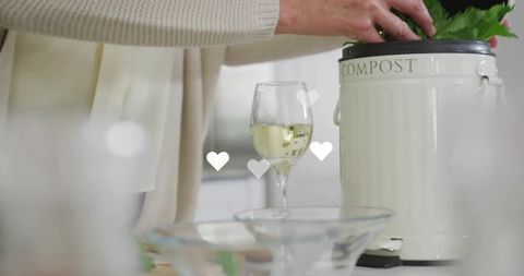 Woman placing kitchen scraps into cream compost bin next to wine glass